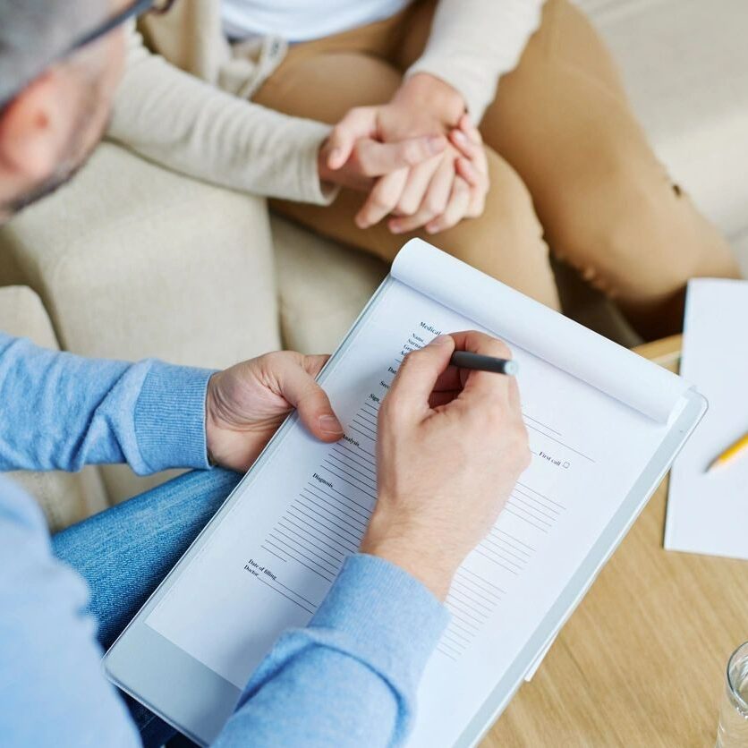 A person writing on paper while another person sits at the table.